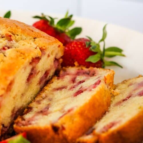 A close-up of sliced strawberry bread from our favorite Strawberry Bread Recipe, with visible strawberry pieces, served on a white plate and garnished with whole strawberries.