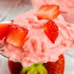 A spoonful of pink strawberry mousse reminiscent of a classic cottage cheese jello salad, topped with small strawberry pieces, with whole strawberries in the background.