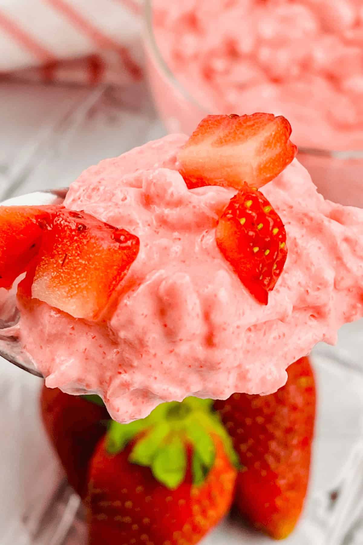 A spoonful of pink strawberry mousse reminiscent of a classic cottage cheese jello salad, topped with small strawberry pieces, with whole strawberries in the background.