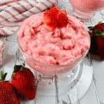 Two glass bowls of creamy pink strawberry dessert, reminiscent of a Cottage Cheese Jello Salad, are topped with strawberry pieces. They sit on a white wooden surface with whole strawberries and a striped cloth nearby.