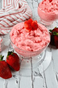 Two glass bowls of creamy pink strawberry dessert, reminiscent of a Cottage Cheese Jello Salad, are topped with strawberry pieces. They sit on a white wooden surface with whole strawberries and a striped cloth nearby.