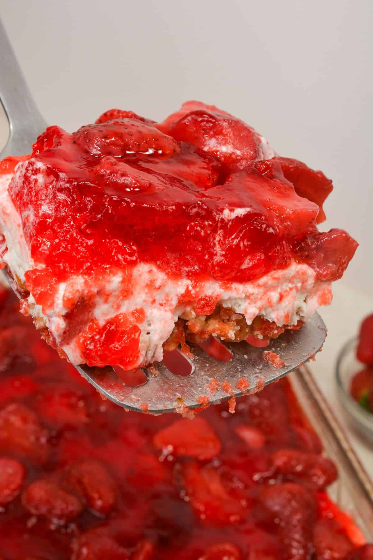 A close-up of a spatula lifting a slice featuring a crumbly pretzel crust, creamy layer, and glossy strawberry topping.