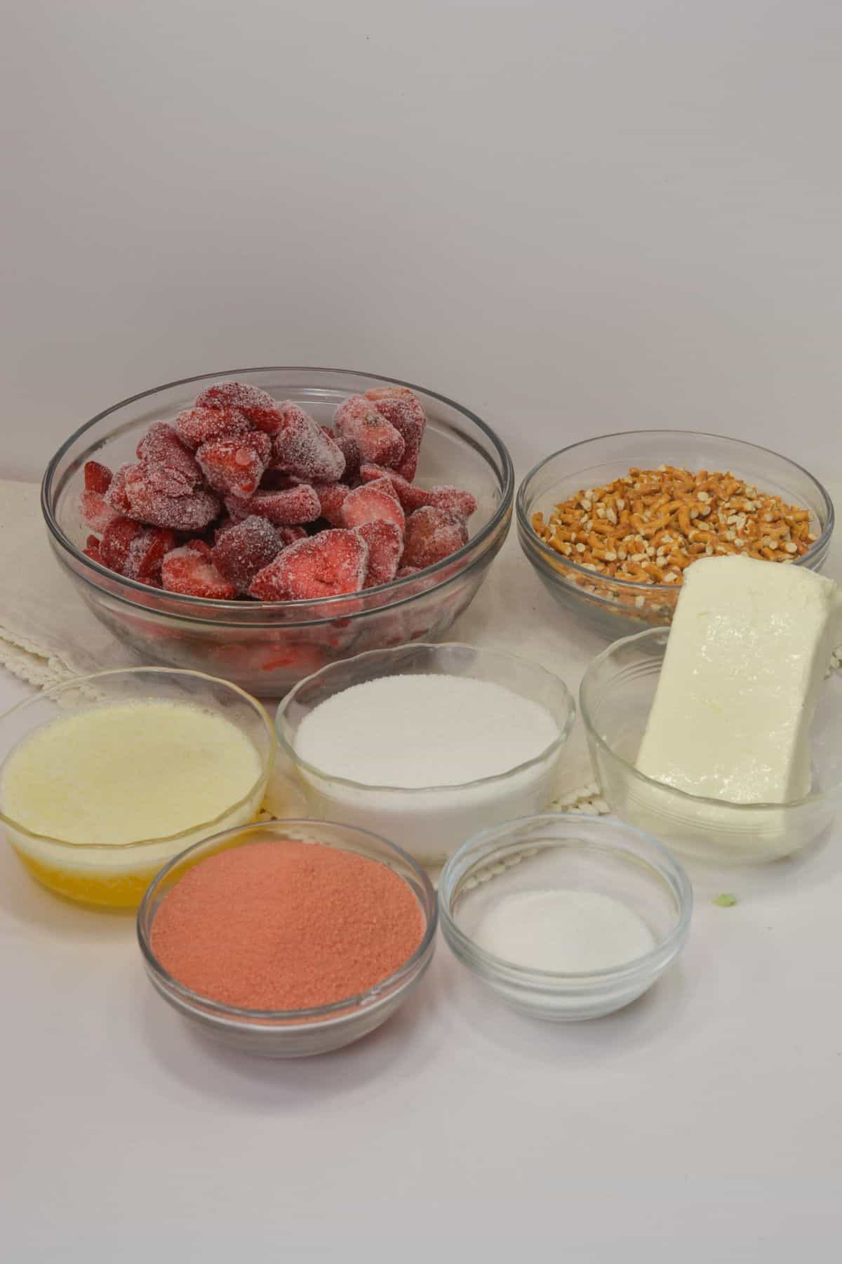 Bowls of frozen strawberries, chopped nuts, melted butter, sugar, powdered strawberry mix, cream cheese, and a small bowl of granulated sugar arranged on a white surface.