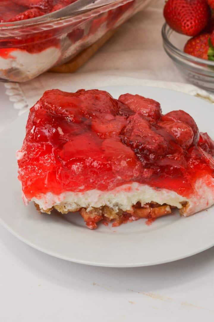A slice of Pretzel Jello Salad with a crumbly base, creamy middle layer, and bright strawberry topping sits on a white plate.