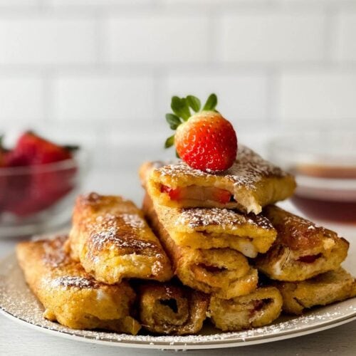 A plate of recipe filled with fruit, dusted with powdered sugar, and topped with a fresh strawberry. A bowl of strawberries and a dipping sauce are in the background.