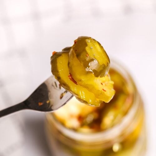 A fork holds up a slice of sweet and spicy pickle above an open jar of pickles, with a blurred checkered cloth in the background.