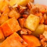 A close-up of a Sweet Potato Apple Casserole bowl, filled with glazed sweet potatoes, apples, pecans, and spices, showing a mix of orange and yellow cubes with a glossy coating.
