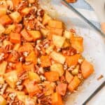 A Sweet Potato Apple Casserole fills a glass baking dish with chopped sweet potatoes, apples, and pecans in a light brown sauce, resting on the countertop beside whole pecans and a spatula.