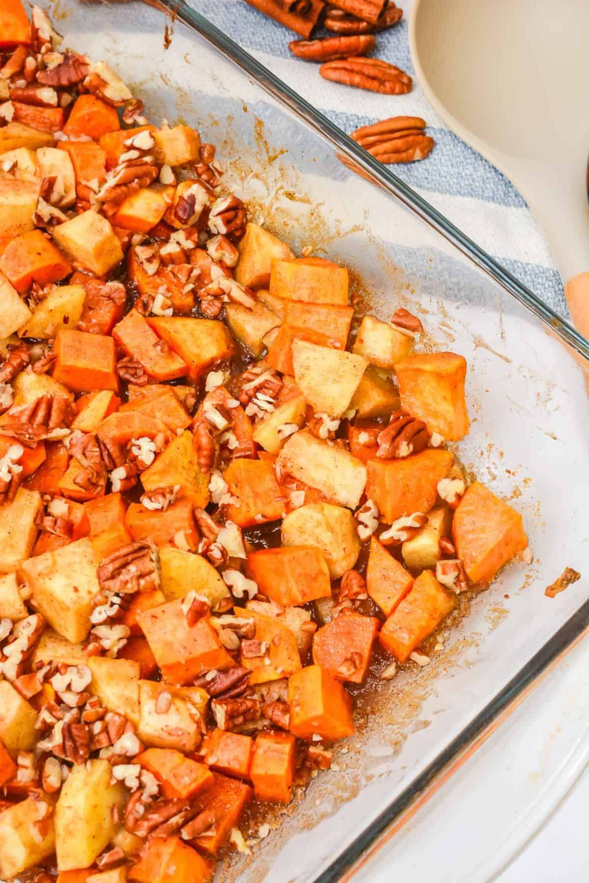 A Sweet Potato Apple Casserole fills a glass baking dish with chopped sweet potatoes, apples, and pecans in a light brown sauce, resting on the countertop beside whole pecans and a spatula.