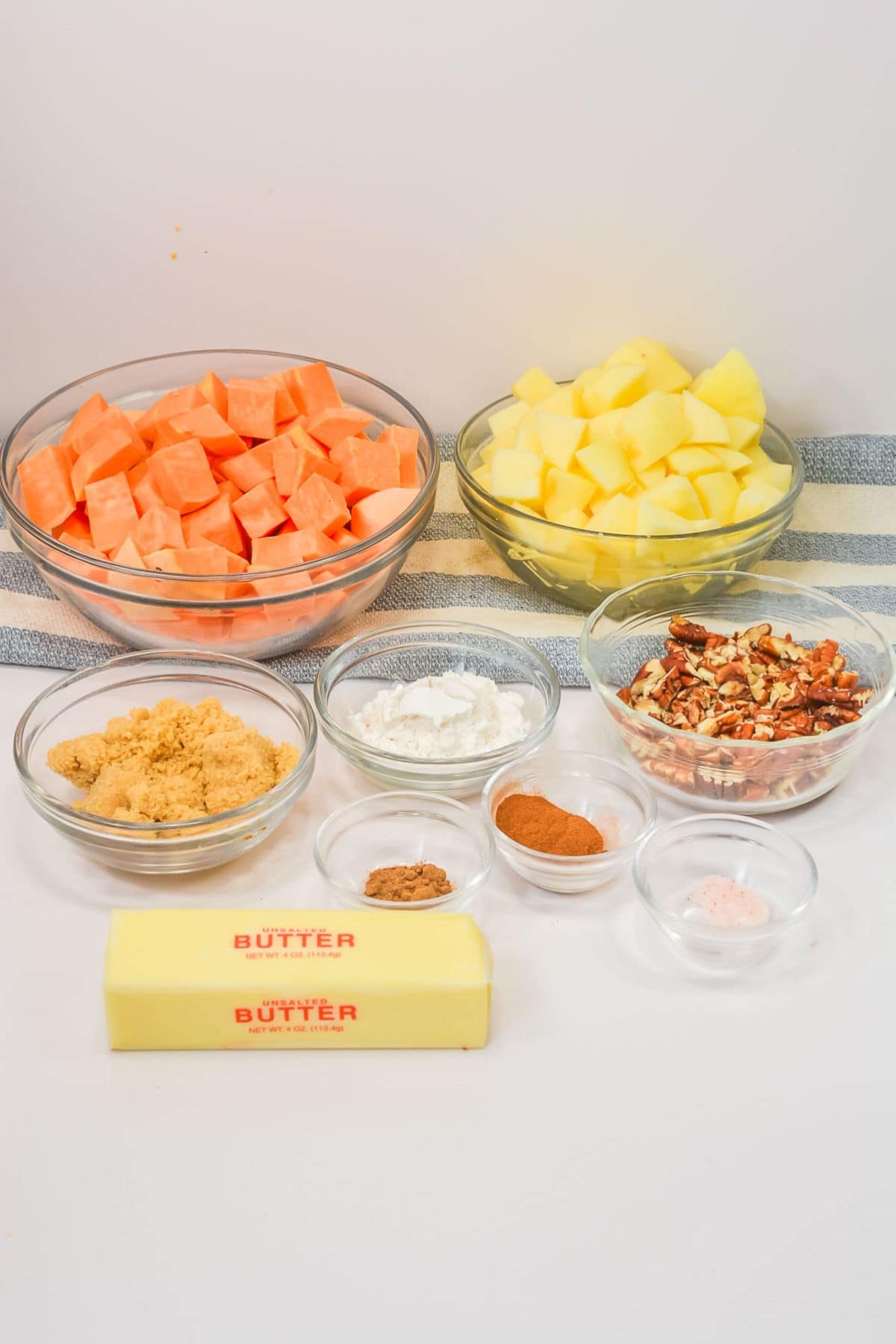Assorted ingredients in glass bowls and on a counter including cubed potatoes, cubed fruit brown sugar, butter, spices, chopped pecans, and salt.