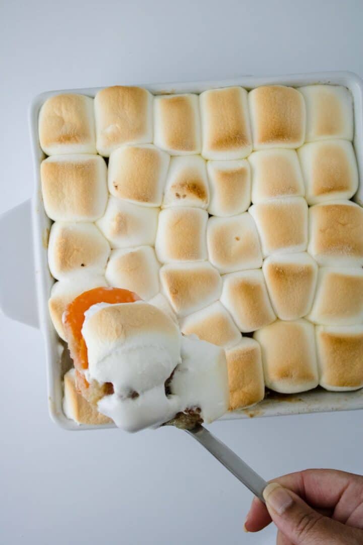 A hand holds a serving spoon with a portion of Sweet Potato Crunch casserole, topped with toasted marshmallows, taken from a baking dish.