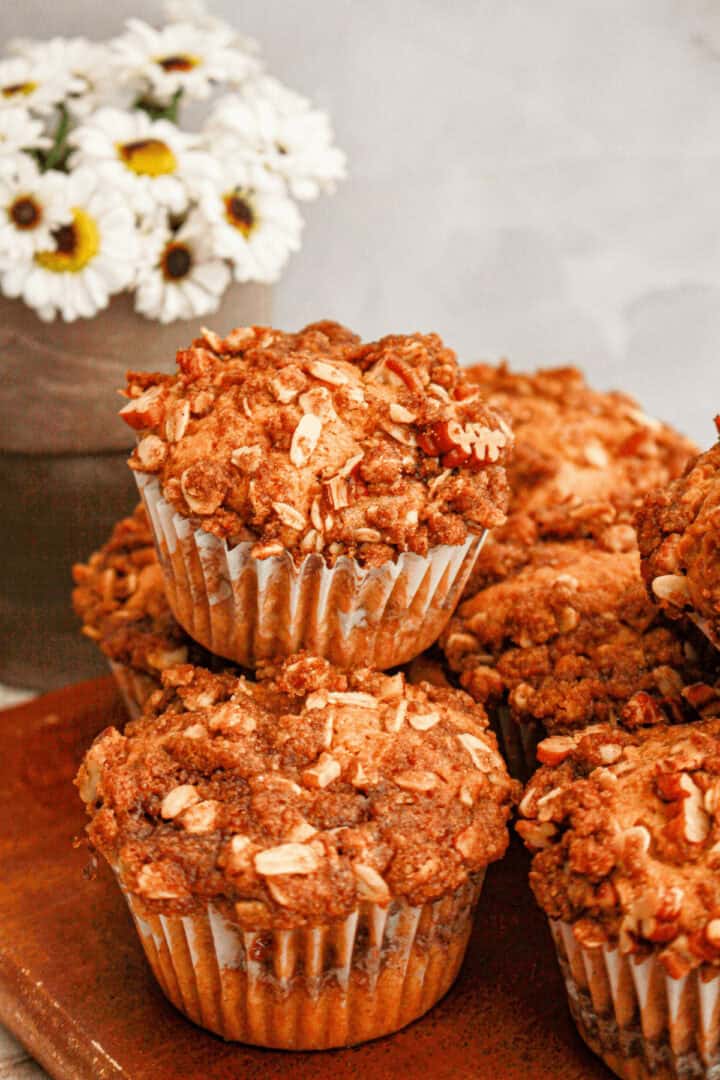 A stack of sweet potato muffins topped with chopped nuts sits on a wooden surface, with a bouquet of white daisies gently complementing the scene in the background.