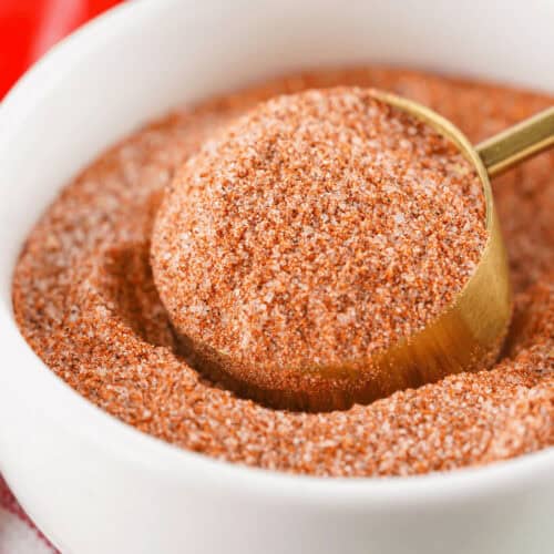 A close-up of a bowl filled with reddish-brown chili lime seasoning. A gold-colored scoop is lifting some of the spice out of the bowl.