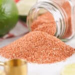 A tipped-over glass jar of seasoning spills onto a white surface next to a vibrant green lime and brass measuring spoons.