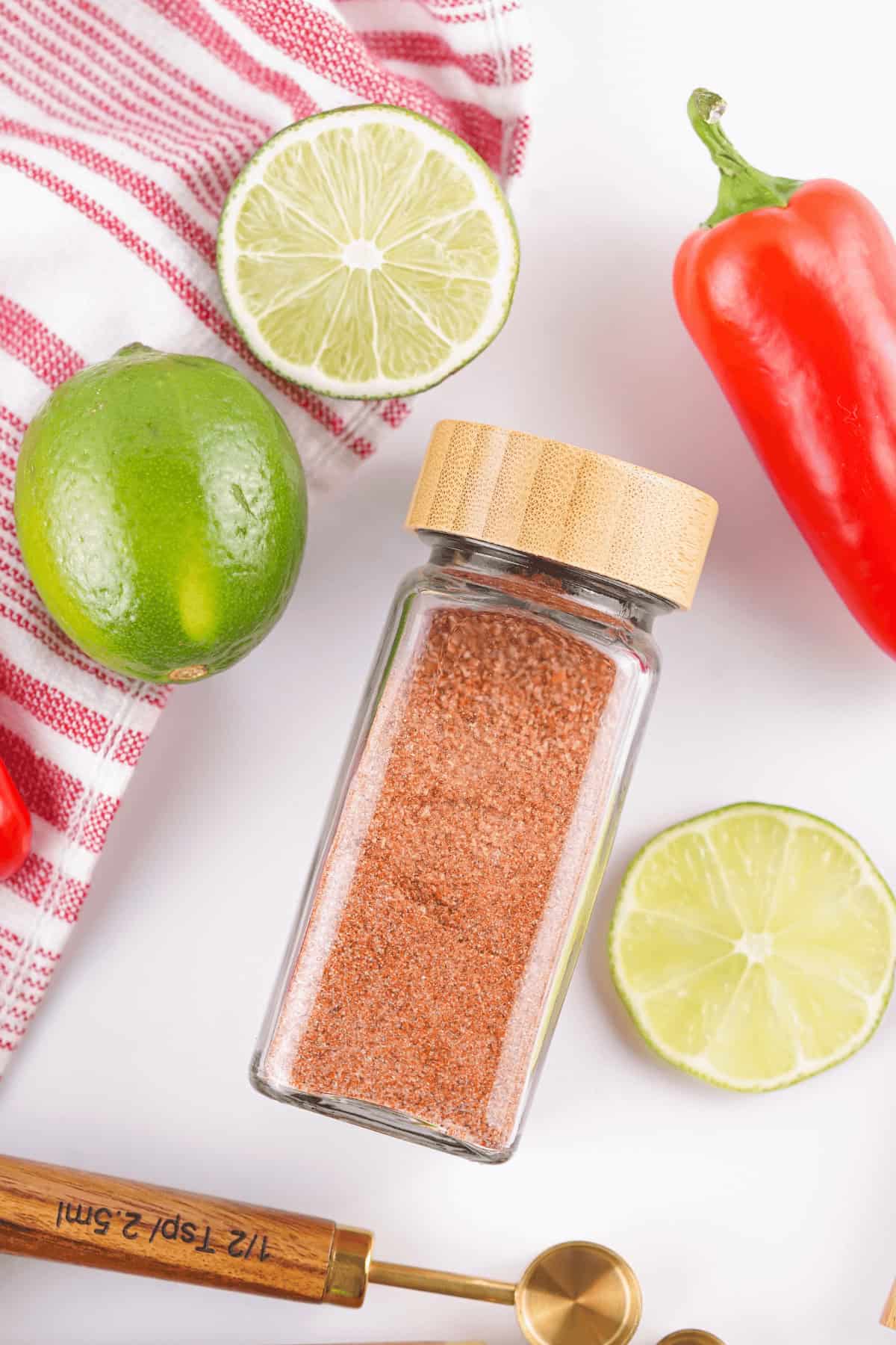 Glass jar with a wooden lid containing Chili Lime Seasoning surrounded by lime slices, a whole lime, a red bell pepper, a red kitchen towel, and measuring spoons.