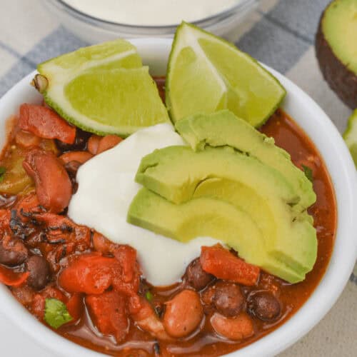 A bowl of three bean chili, topped with sliced avocado, lime wedges, and a dollop of sour cream. A bowl of sour cream and half an avocado are in the background.