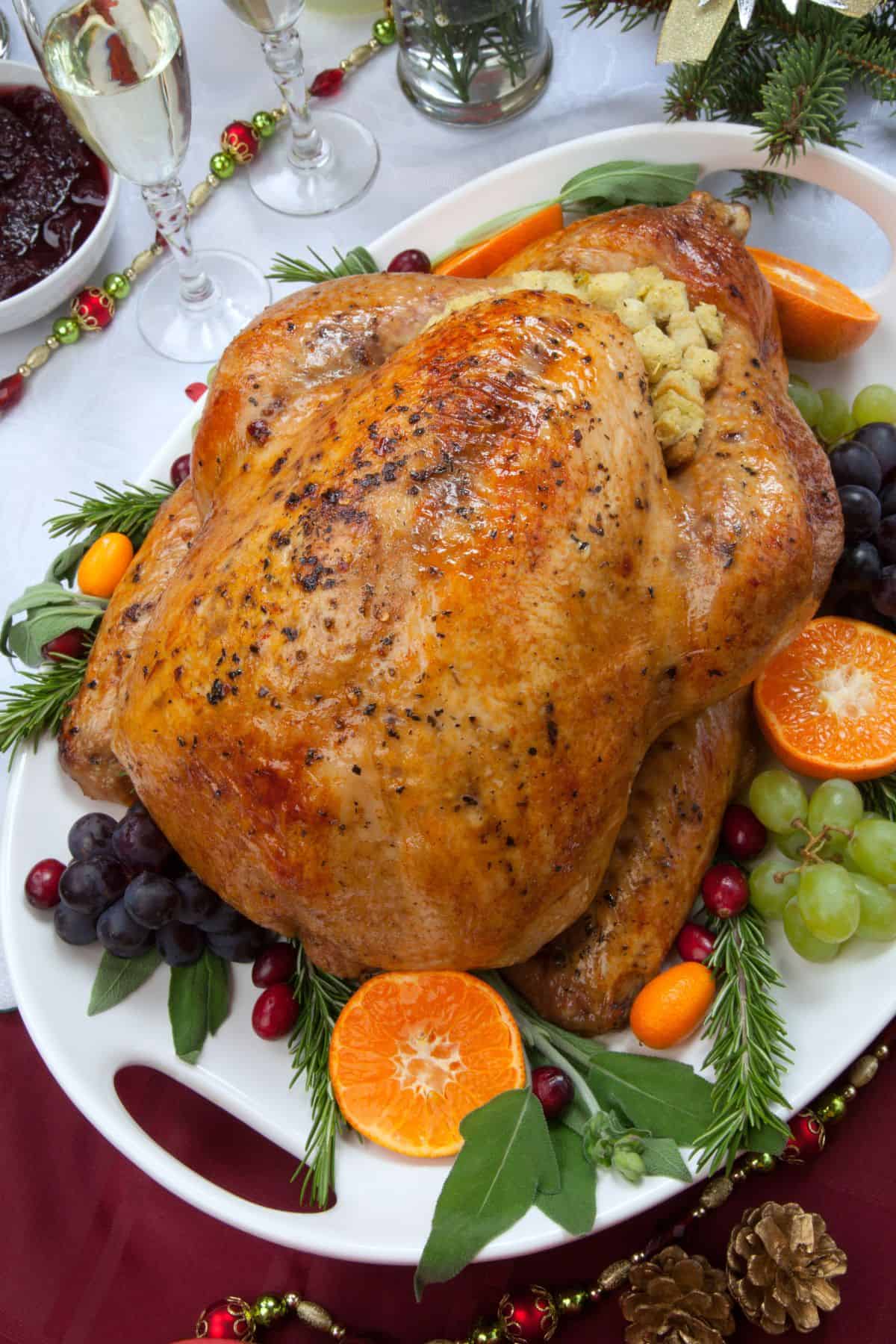 A Turkey in an Oven Bag stuffed with bread stuffing is served on a platter with grapes, cranberries, orange slices, carrots, and herbs on a decorated table.