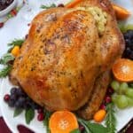 A roasted, stuffed turkey in an oven bag is served on a white platter with assorted fruits, vegetables, and herbs, set on a festive table with wine glasses and holiday decorations.
