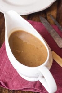 A white gravy boat filled with Turkey Gravy from the Drippings sits on a red cloth napkin, next to a plate, knife, and fork on a wooden table.