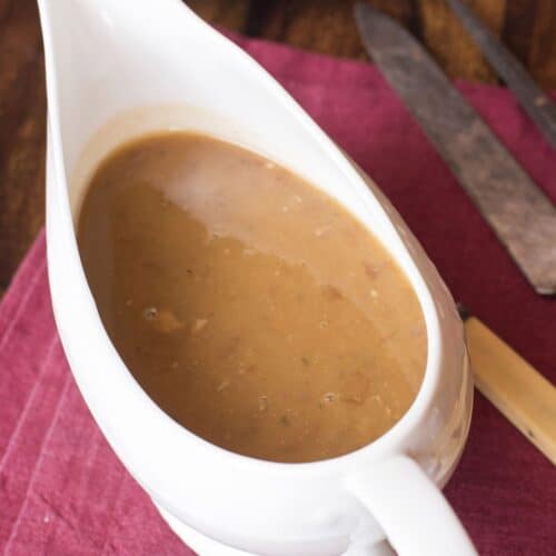 A white gravy boat filled with Turkey Gravy from the Drippings sits on a red cloth napkin, next to a plate, knife, and fork on a wooden table.