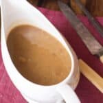 A white gravy boat filled with rich Turkey Gravy from the Drippings sits on a red cloth napkin next to a knife and fork on a wooden table.