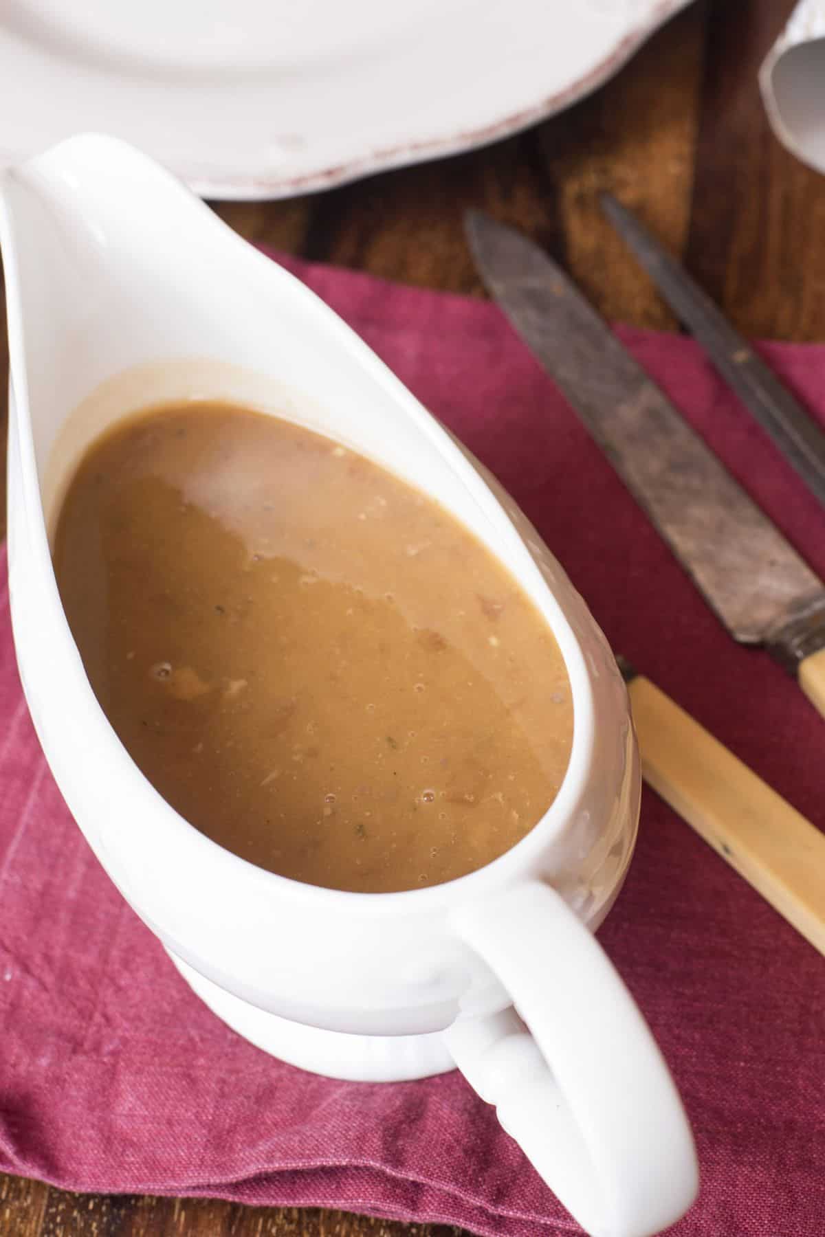 A white gravy boat filled with rich Turkey Gravy from the Drippings sits on a red cloth napkin next to a knife and fork on a wooden table.