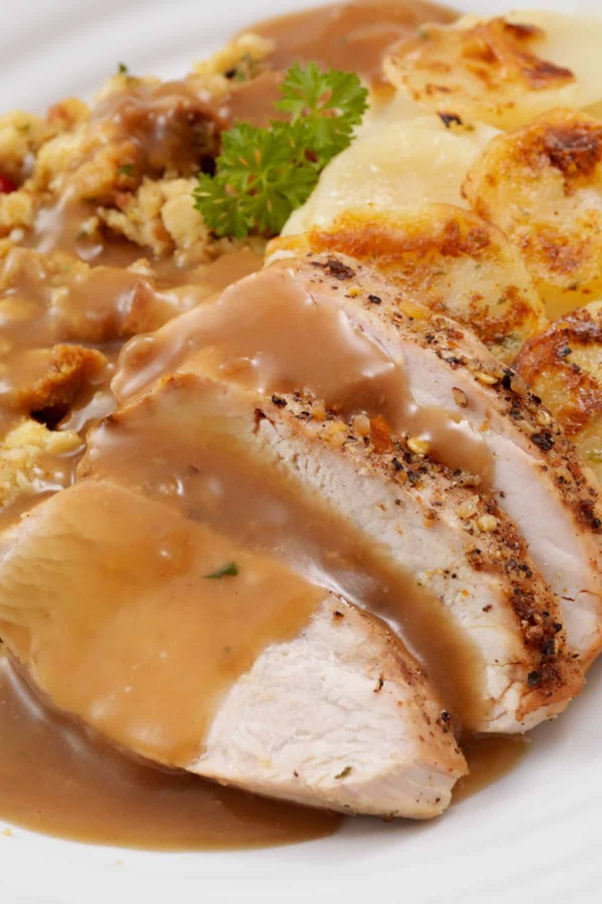 Sliced roast turkey with Turkey Gravy from the Drippings, scalloped potatoes, stuffing, and a parsley garnish on a white plate.