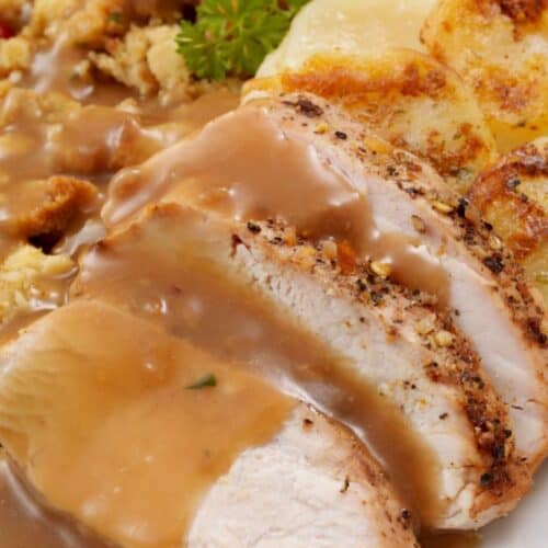 Sliced roasted turkey breast with Turkey Gravy from the Drippings, served alongside scalloped potatoes and stuffing, garnished with a parsley sprig on a white plate.