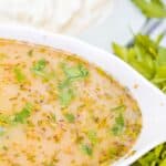 A white oval dish filled with a light brown, herb-flecked Turkey Gravy without Turkey Drippings, garnished with chopped parsley, sits beside fresh herbs and a blurred plate of rice in the background.