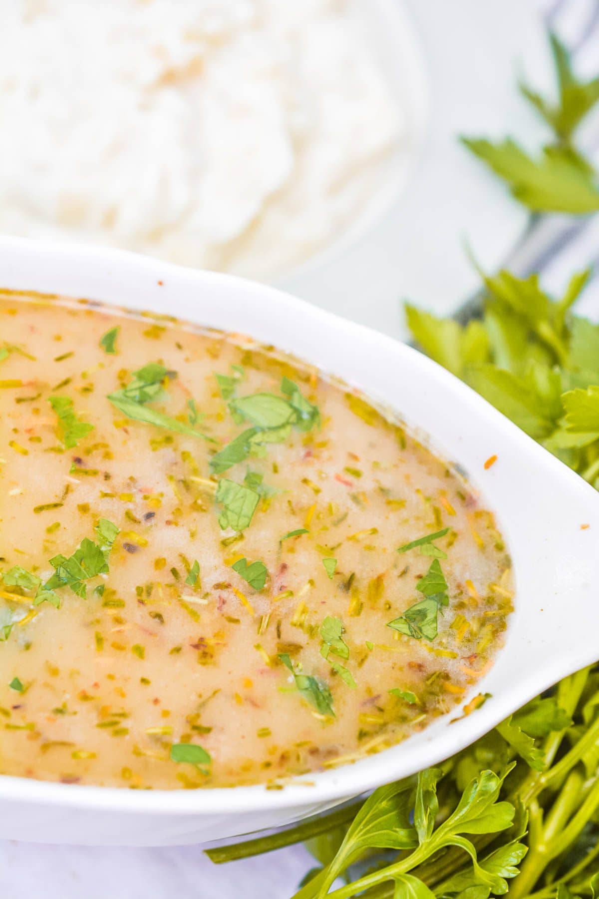 A white oval dish filled with a light brown, herb-flecked Turkey Gravy without Turkey Drippings, garnished with chopped parsley, sits beside fresh herbs and a blurred plate of rice in the background.
