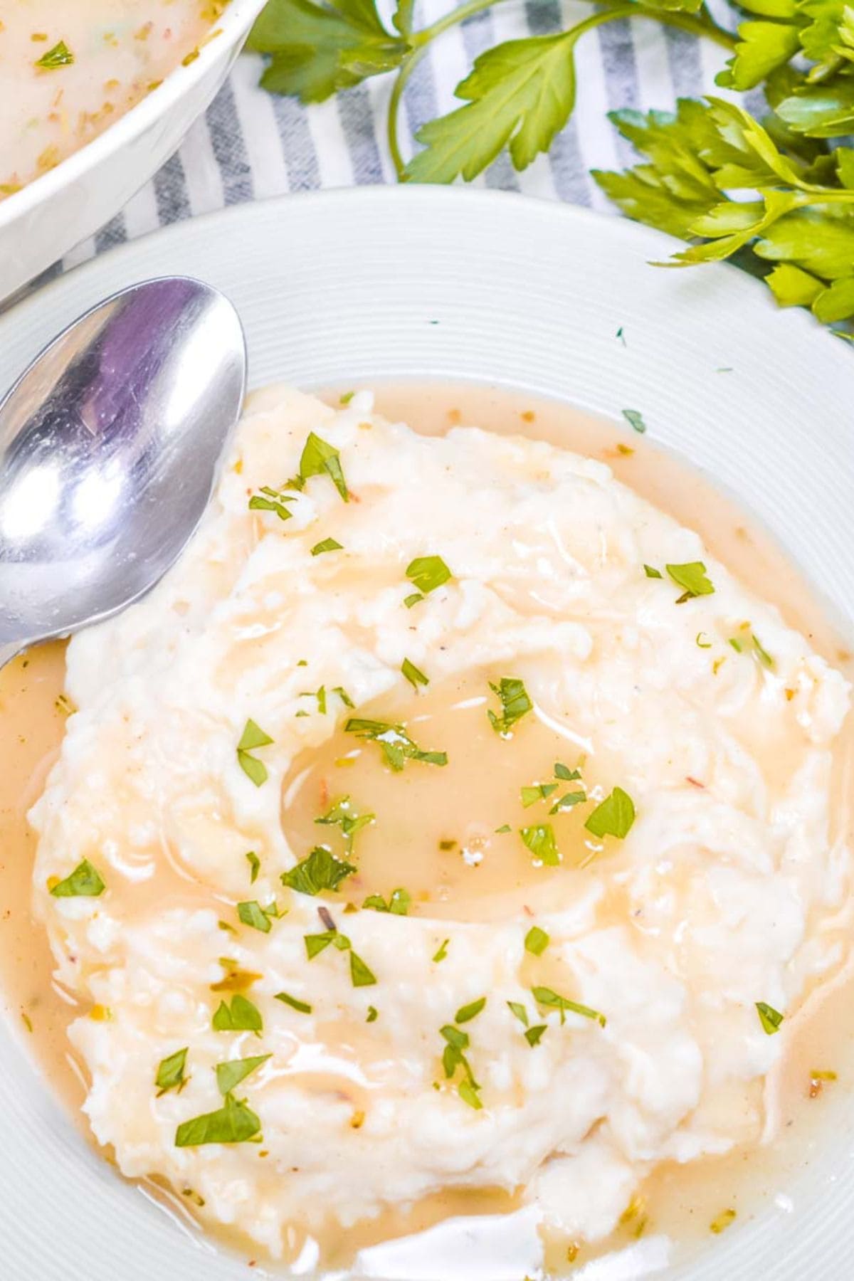 A plate of mashed potatoes topped with  and chopped parsley, next to a spoon and fresh parsley on a striped cloth.