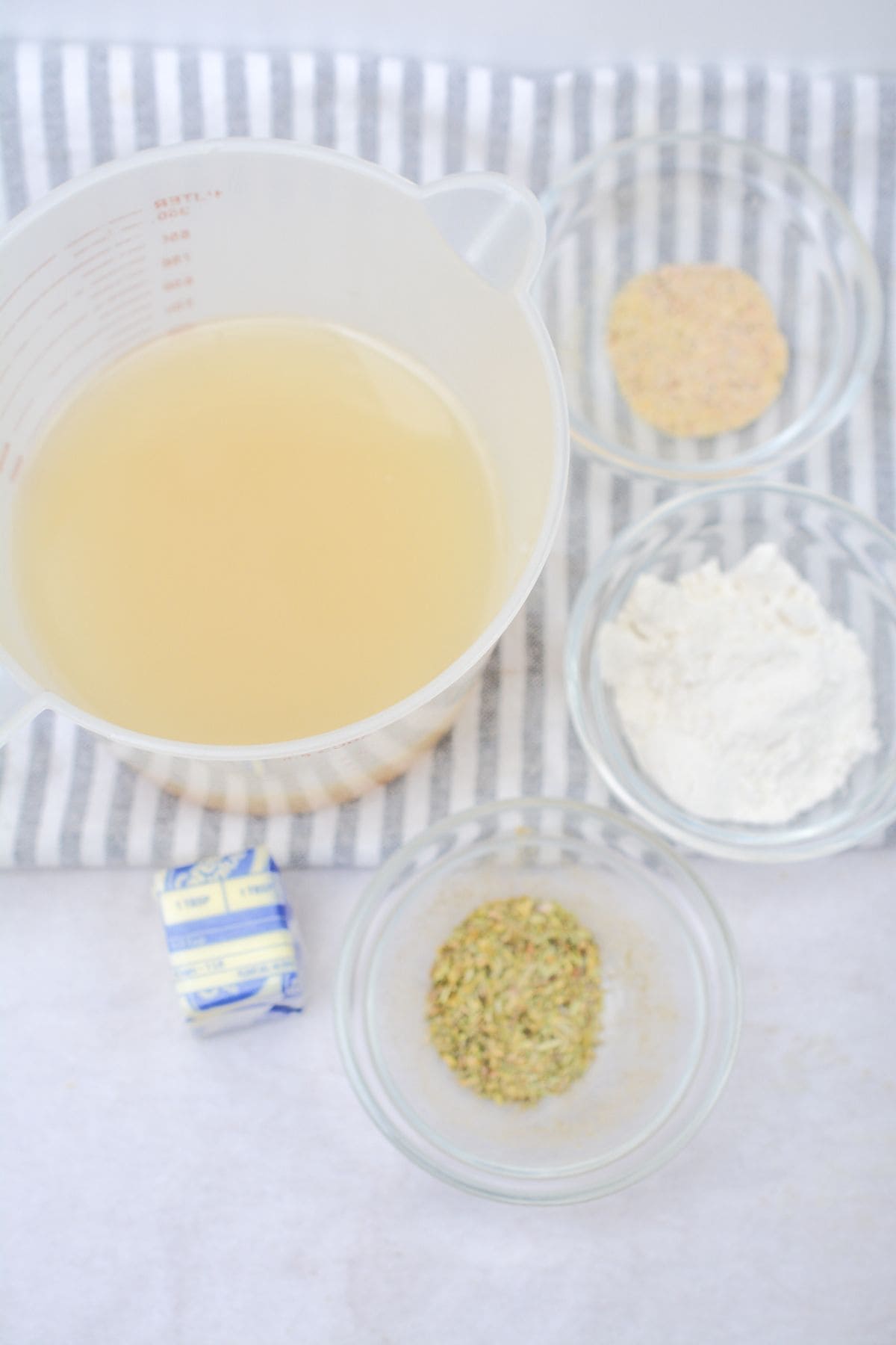 A measuring cup of broth, three small bowls with flour, crushed seasoning, and another spice blend, plus a wrapped cube of butter are arranged on a striped cloth.