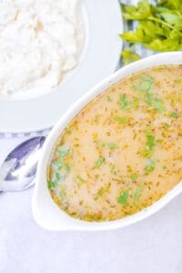 A white dish of Turkey Gravy without Turkey Drippings, garnished with chopped herbs, sits next to a plate of mashed potatoes and a bunch of fresh parsley.
