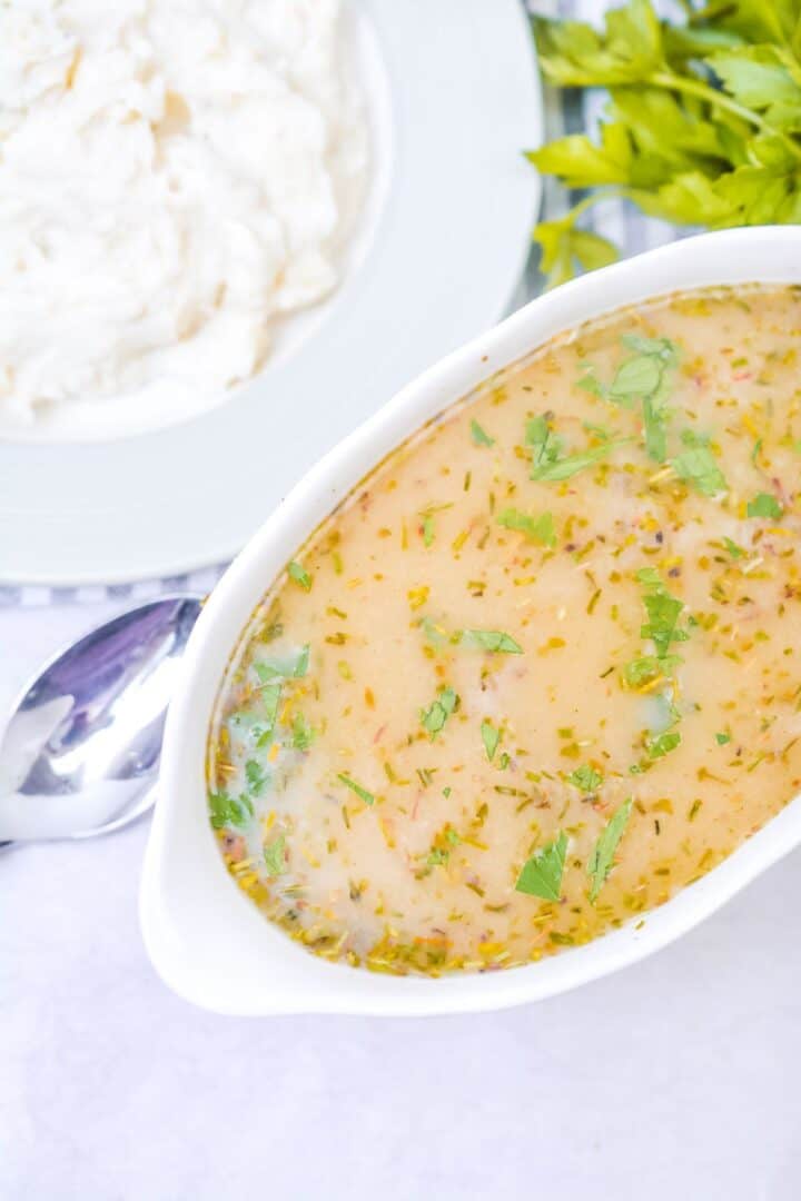 A white dish of Turkey Gravy without Turkey Drippings, garnished with chopped herbs, sits next to a plate of mashed potatoes and a bunch of fresh parsley.