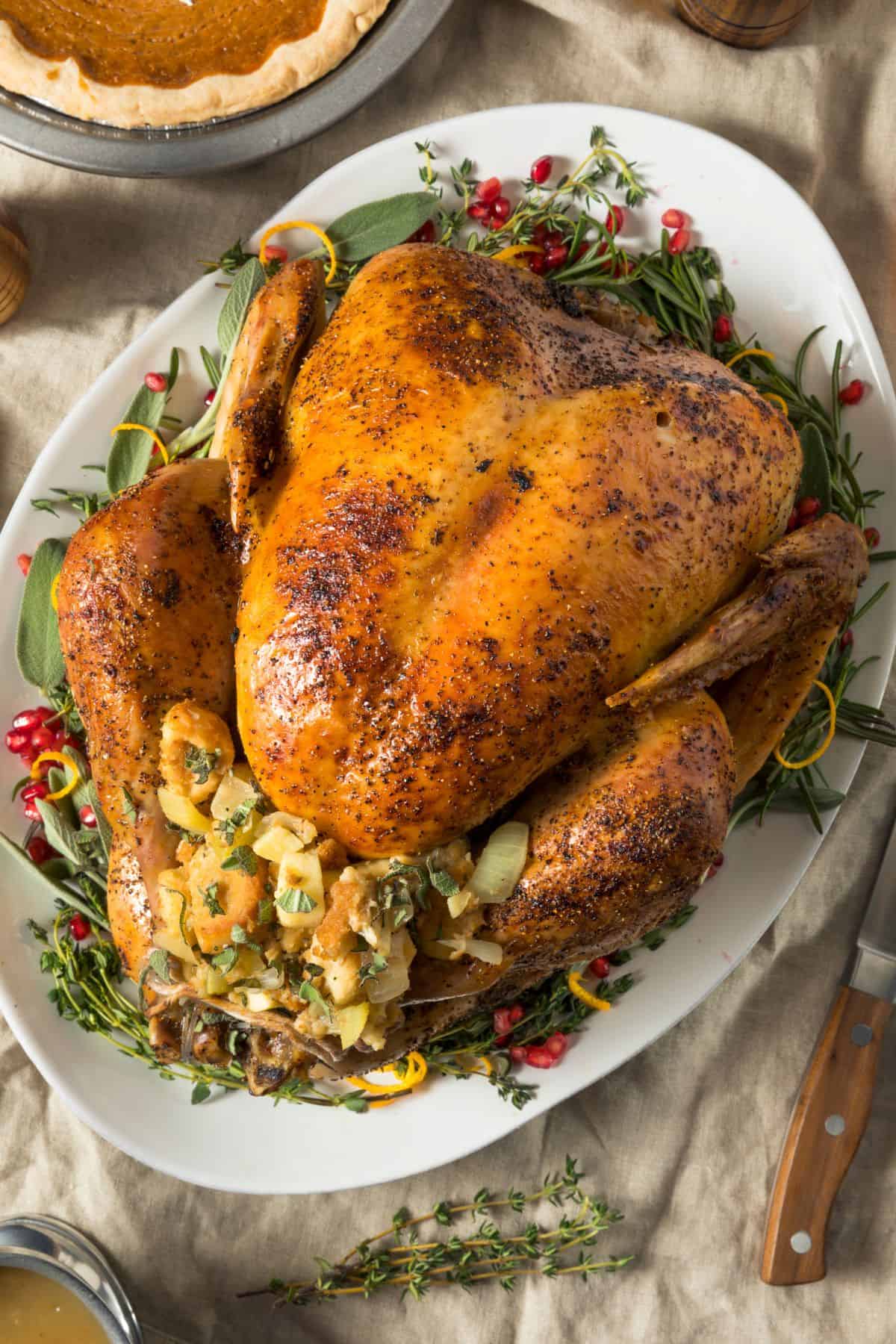 Roast turkey in the roaster oven with golden skin, stuffed with bread and herbs, served on a white platter with fresh greens, with a pie partially visible in the background.