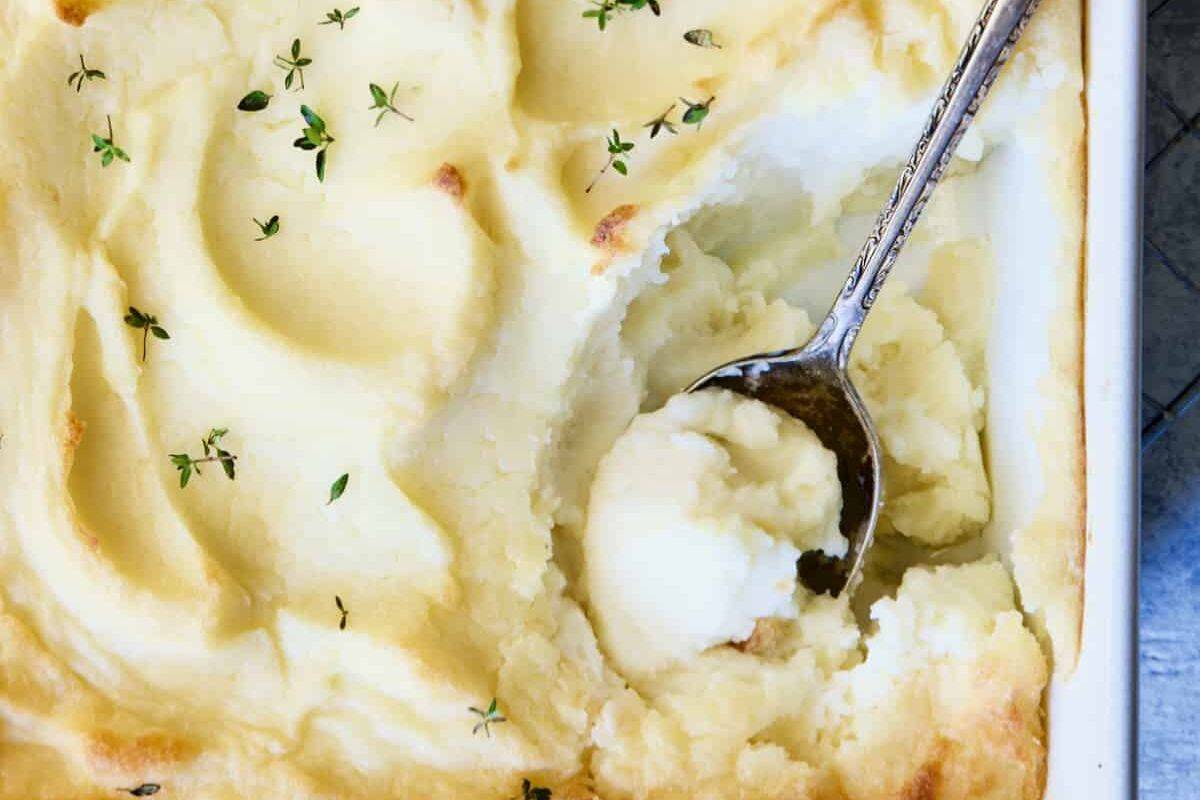 A serving spoon scoops creamy mashed potatoes from a baking dish, garnished with fresh thyme leaves—perfect for easy appetizers at your next gathering.