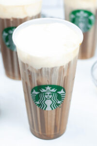A transparent cup with Vanilla Sweet Cream Cold Brew topped with creamy froth, featuring a Starbucks logo. Three similar cups are partially visible in the background.