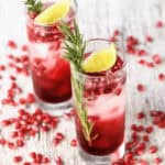Two glasses of pomegranate cocktail with lime slices and rosemary sprigs on a rustic wooden surface, scattered with pomegranate seeds.