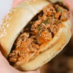 A close-up of two hands holding a sandwich with a soft bun and a filling of seasoned ground meat mixture.