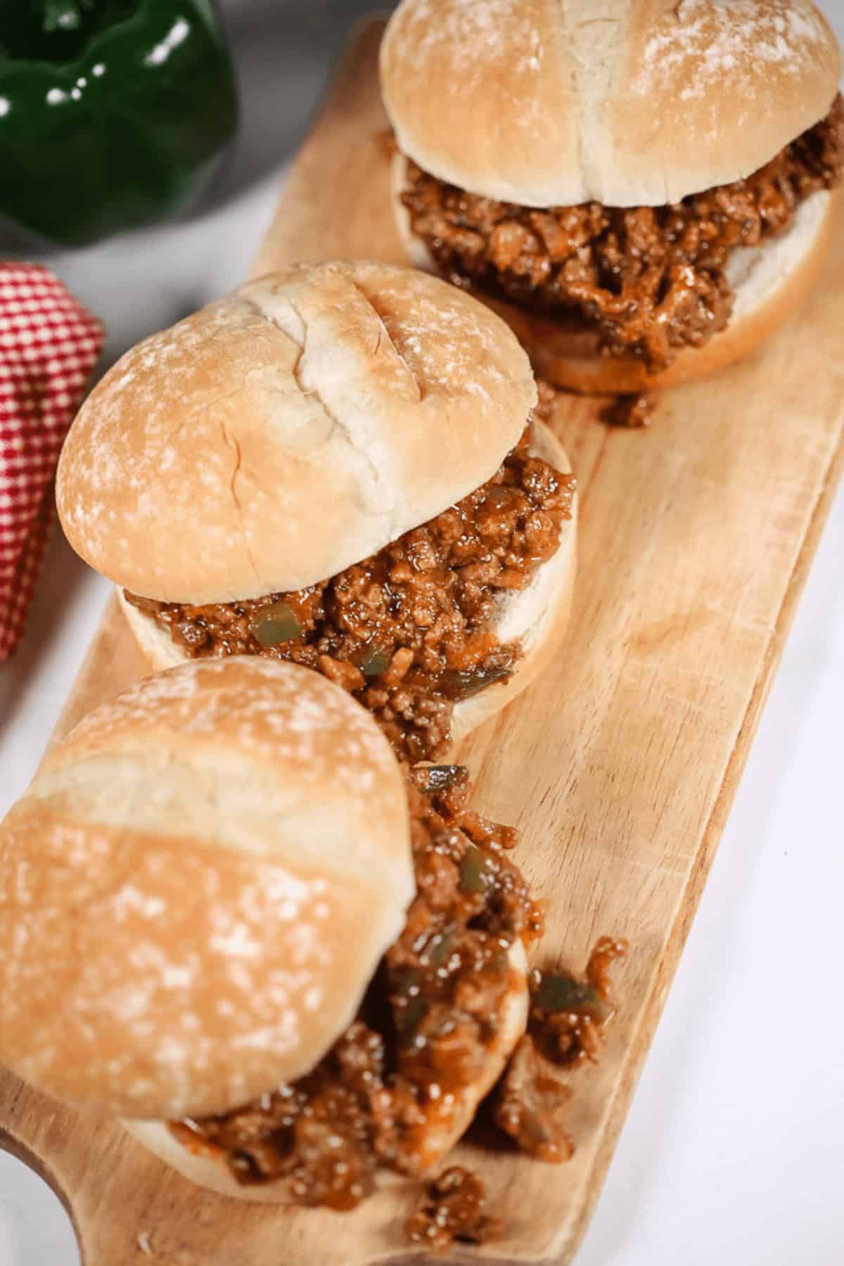 Three Wimpies (Easy Sloppy Joes) with ground meat filling on a wooden cutting board. A red checkered cloth and a green pepper are visible in the background.