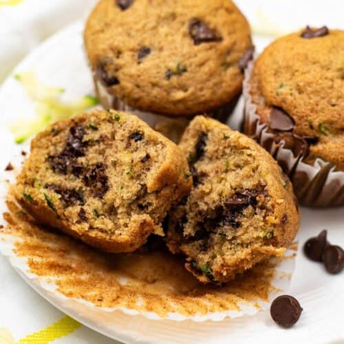 Three Zucchini Chocolate Chip Muffins sit on a white plate, with one split open to reveal its moist interior filled with chocolate chips and green zucchini pieces.