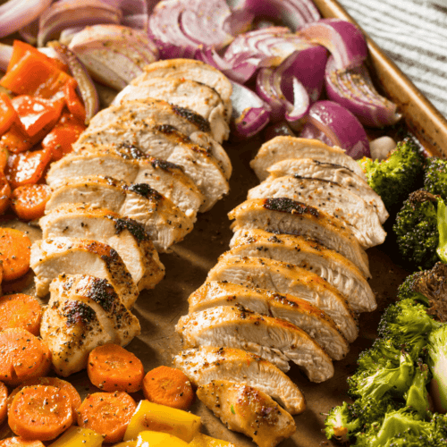 A balsamic chicken and vegetable sheet pan features slices of grilled chicken breast surrounded by roasted vegetables, including red onions, bell peppers, carrots, and broccoli.