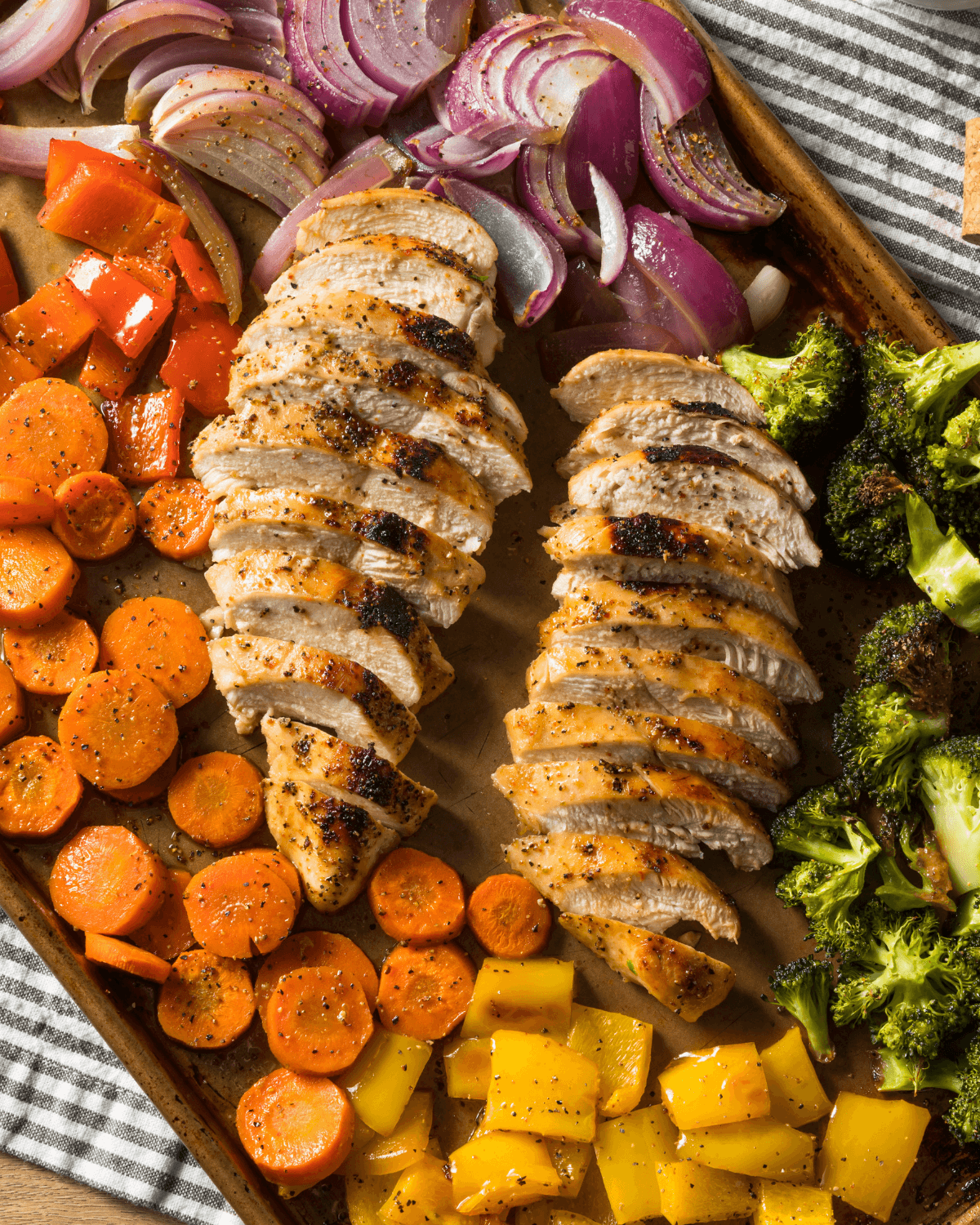 A tray of Balsamic Chicken and Vegetable Sheet Pan features sliced roasted chicken breasts surrounded by seasoned vegetables including broccoli, red onions, carrots, yellow bell peppers, and red bell peppers.
