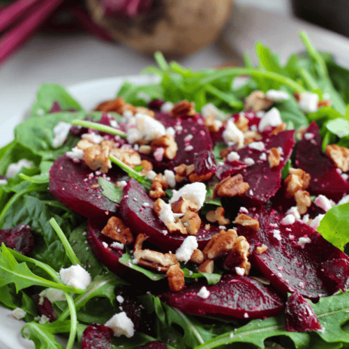 A fresh beet salad with feta features arugula, sliced beets, crumbled feta cheese, and chopped pecans on a white plate. A whole beet and greens are in the background.