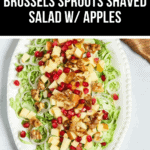 A plate of Brussels Sprouts Shaved Salad is beautifully topped with apples, walnuts, and pomegranate seeds. A banner above proudly declares it a "Fall Favorite: Brussels Sprouts Shaved Salad w/ Apples.