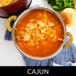 A bowl of flavorful Cajun Chicken and Rice soup sits on a cloth, with a pot in the background. It's garnished with fresh parsley, while an onion rests nearby, enhancing its vibrant appeal.