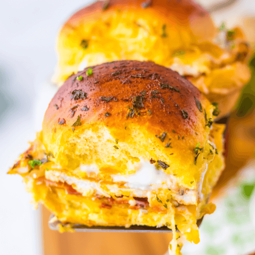 A close-up of a golden-brown Chicken Bacon Ranch Slider filled with gooey melted cheese and herbs, held by a spatula.