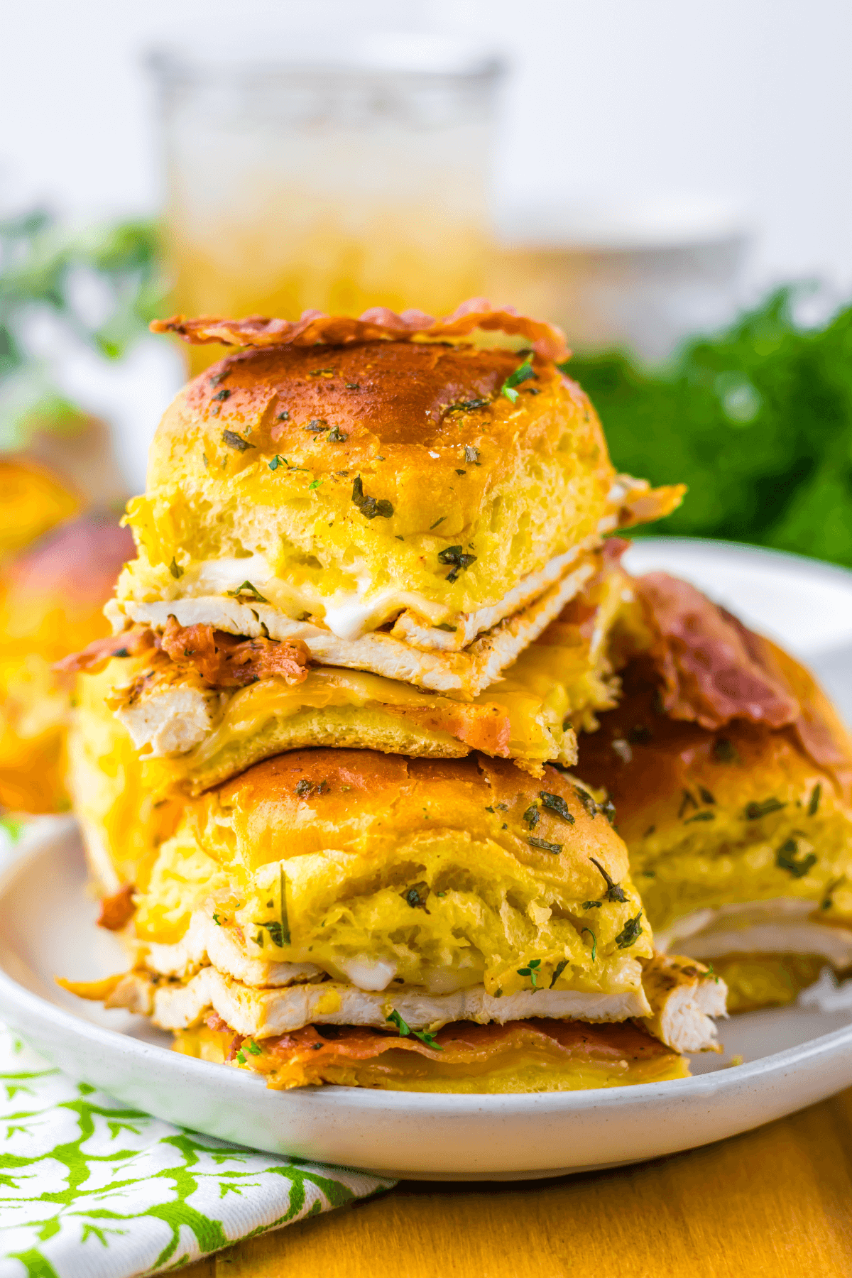 Two stacks of sandwiches with melted cheese on fluffy buns are garnished with herbs and served on a white plate. A green napkin and a drink are in the background.