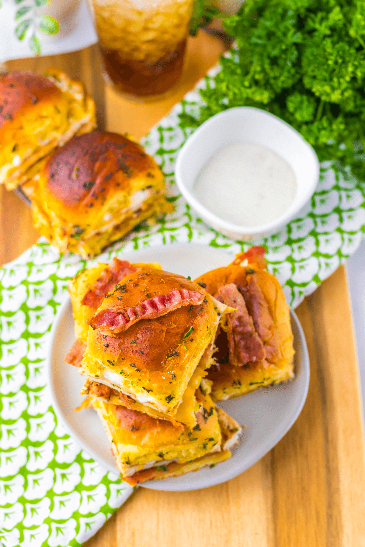 Plate of stacked Chicken Bacon Ranch Sliders on a wooden table, garnished with fresh parsley and served with a bowl of dipping sauce and a glass of soda in the background.