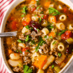A bowl of hearty vegetable and beef soup with pasta, reminiscent of Olive Garden's Pasta Fagioli, garnished with parsley, rests beside a red and white striped cloth.